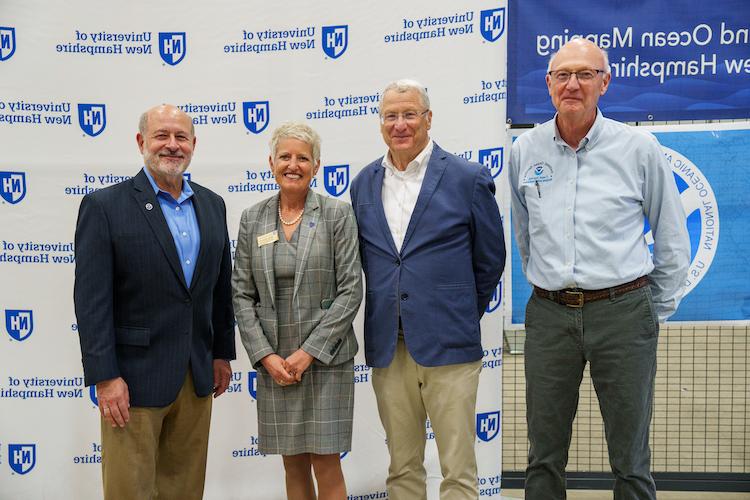 Four people stand in front of a UNH banner