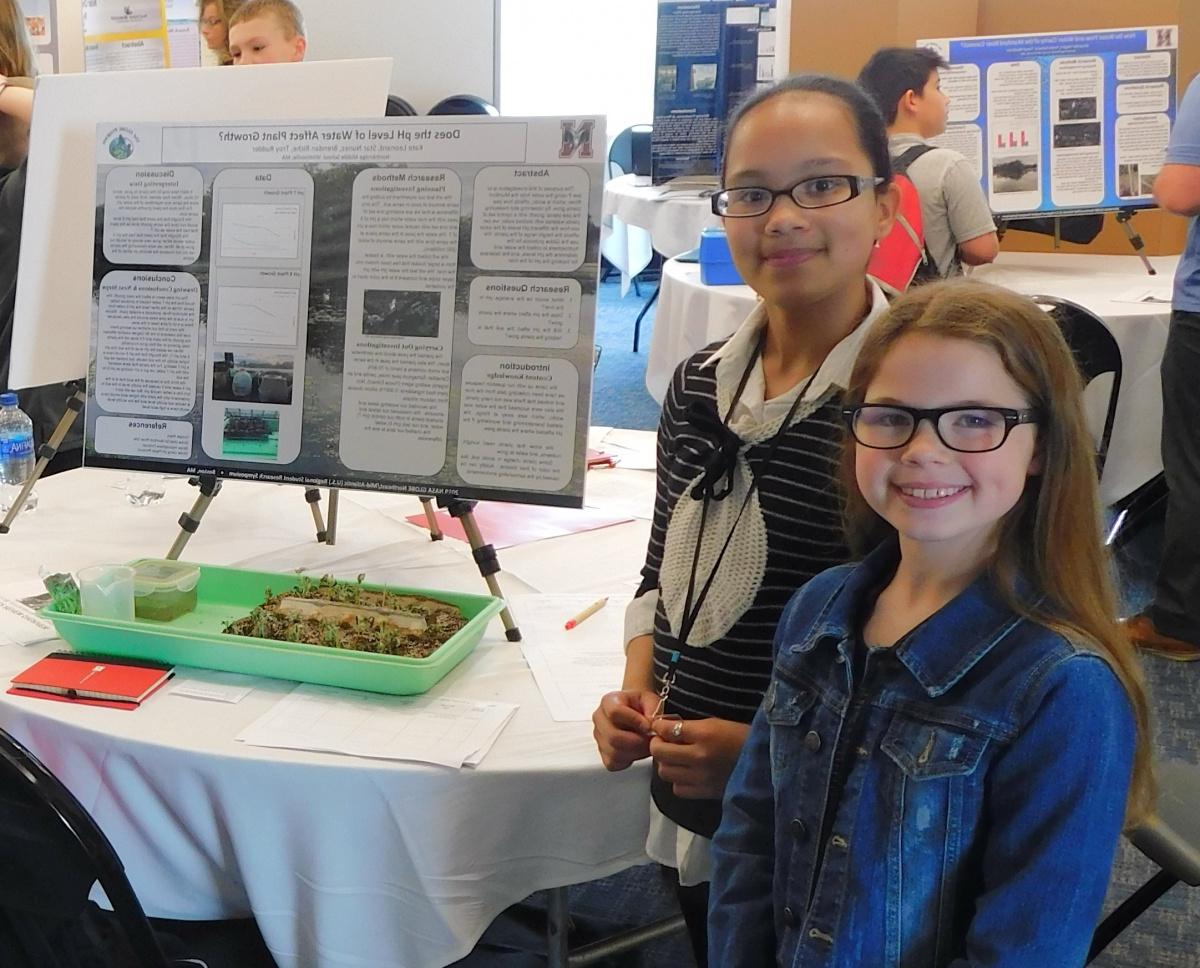 Two middle school girls st和 beside a scientific poster, smiling